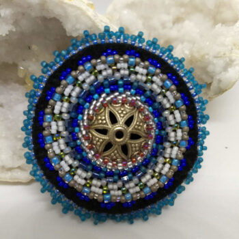 Sinised ja valged helmed tikitud Mandala prossile. Keskel on metalne südamik. Mandala on loodud helmestikandina. heledal taustal.