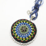 värviliste helmestega tikitud EESTI teemaline Mandala kaelakee. Keskel on metalne sõle südamik. Mandala on loodud helmestikandina. Sinine pael. heledal taustal.