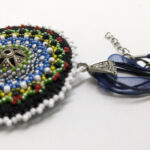 värviliste helmestega tikitud EESTI teemaline Mandala kaelakee. Keskel on metalne sõle südamik. Mandala on loodud helmestikandina. Sinine pael. heledal taustal.