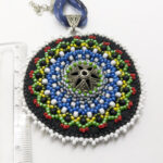 värviliste helmestega tikitud EESTI teemaline Mandala kaelakee. Keskel on metalne sõle südamik. Mandala on loodud helmestikandina. Sinine pael. heledal taustal.