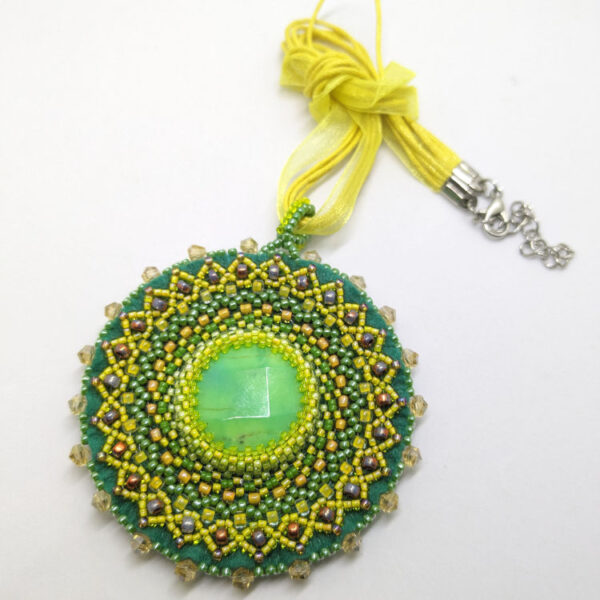 Rohelised ja kollased helmed tikitud Mandala kaelakee. Keskel on KRÜSIPRAAS kristallist südamik. Mandala on loodud helmestikandina. kollane pael. heledal taustal.